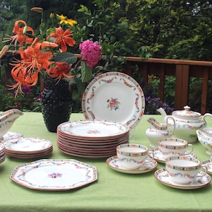 May include: A complete vintage porcelain tea set with floral and ribbon designs. The set includes plates, cups, saucers, a teapot, creamer, sugar bowl, and a gravy boat. The set is arranged on a green tablecloth with a vase of flowers.