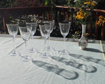 May include: A set of six clear glass wine glasses with silver rims sit on a white tablecloth. The glasses are arranged in a semi-circle, with the stems pointing towards the center. The glasses are empty and reflect the light from the window.