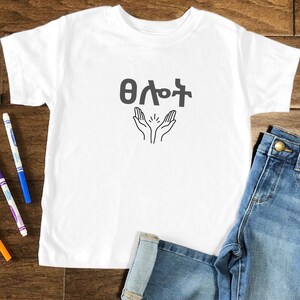 May include: White t-shirt with the Amharic phrase "ሰላም" and a graphic of two hands raised in prayer.