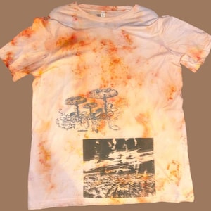 May include: A light-colored t-shirt with a tie-dye effect in shades of orange and yellow. The shirt features a graphic of three mushrooms and a landscape scene. The t-shirt is a crew neck with short sleeves.
