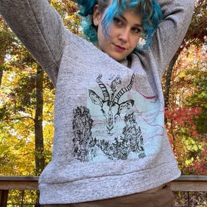 Puede incluir: Una sudadera gris con un estampado gráfico en blanco y negro de una cabeza de antílope con cuernos. El estampado está rodeado de un diseño floral. La sudadera la lleva puesta una persona con el pelo azul.
