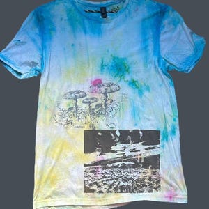 May include: A tie-dye t-shirt with a blue, yellow, and white color scheme. The shirt features a graphic of three mushrooms with floral accents and a black and white landscape scene. The shirt has a crew neck.