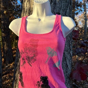 May include: A bright pink ribbed tank top with a square neckline. The tank top features black and gray graphic prints of flowers and frogs. The tank top is displayed on a white mannequin in front of a tree.