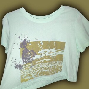 May include: A light green t-shirt featuring a graphic print of a woman in a flowing dress against a landscape. The design is in shades of gold, purple, and gray, with paint splatters around the edges. The shirt has a crew neck and short sleeves.