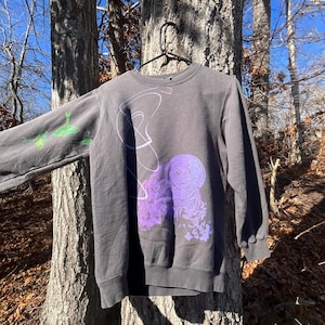 Könnte beinhalten: Dunkelgraues Sweatshirt mit Rundhalsausschnitt und langen Ärmeln. Das Design zeigt eine abstrakte lila und weiße Grafik auf der Vorderseite und grüne und lila Grafiken auf dem linken Ärmel. Das Sweatshirt hängt an einem schwarzen Kleiderbügel.