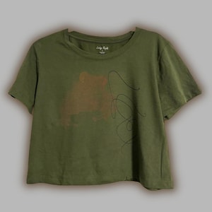May include: Olive green cropped t-shirt with a brown abstract design and black line art. The shirt has a crew neck and short sleeves. The text "Loving People" is visible on the tag.