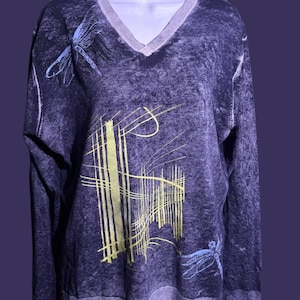 Può includere: Un maglione viola scuro con scollo a V dall'aspetto strutturato. Il maglione presenta un disegno di linee gialle e bianche e due libellule blu. Lo scollo e i polsini sono di un viola più chiaro.