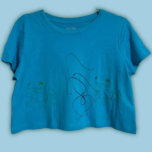 May include: A turquoise crop top with a crew neck. The shirt features a line drawing of two frogs and a curved line design. The frogs have green eyes and are positioned on either side of the curved line.