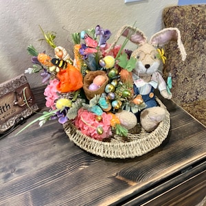 May include: A woven basket filled with a spring-themed floral arrangement. The arrangement features pink, orange, and purple flowers, green leaves, and a small bunny wearing overalls. There are also small carrots, butterflies, and Easter eggs in the basket.