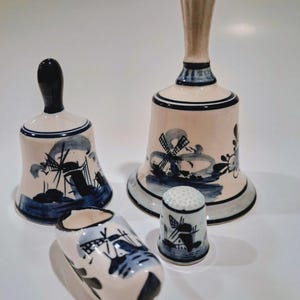 May include: A collection of Dutch Delftware items, including two bells, a wooden shoe, and a thimble. Each piece is painted with a windmill scene in blue and white. The bells have tan handles and rims.