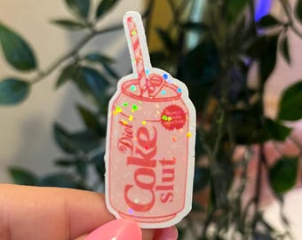 May include: A pink Diet Coke can-shaped sticker with the words "Diet Coke Slut" in white. The sticker has a straw and glitter accents. The background is blurred with green foliage.