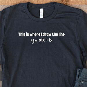 May include: A black t-shirt with white text that reads "This is where I draw the line y=mx+b". The shirt is laid out on a wooden surface with a pair of white sneakers and blue jeans.