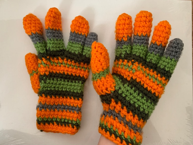 May include: A pair of handmade crochet gloves. The gloves have a striped pattern in orange, green, and grey. The fingers and cuffs are orange. The gloves are designed for warmth and style.