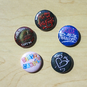 Może przedstawiać: Pięć okrągłych znaczków z różnymi wzorami. Znaczek są czarne, niebieskie, różowe i zielone z białym tekstem. Tekst na znaczkach brzmi "Fig and the Sig Figs", "Julie and the Phantoms", "Dirty Cando", "every place i cry" i "Corroded Coffin".