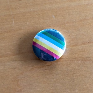 May include: A round button with a colorful striped design. The button features diagonal stripes in shades of blue, green, white, yellow, and pink. The button is set against a light brown wooden surface.