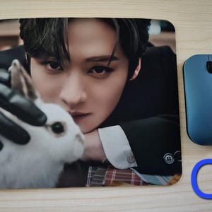 May include: Rectangular mouse mat featuring a close-up image of a person with dark hair and a white rabbit. A blue wireless mouse is to the right. The mouse mat has a smooth surface.