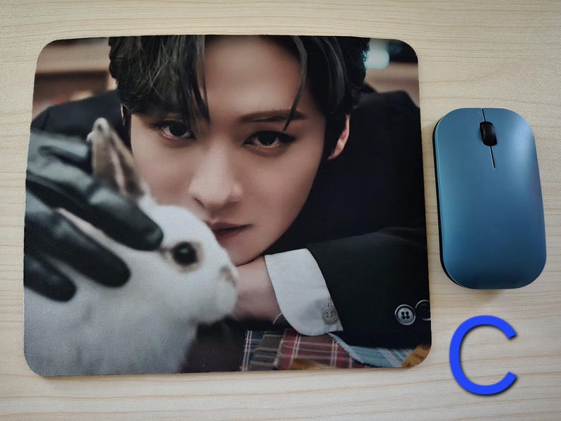 May include: Rectangular mouse mat featuring a close-up image of a person with dark hair and a white rabbit. A blue wireless mouse is to the right. The mouse mat has a smooth surface.