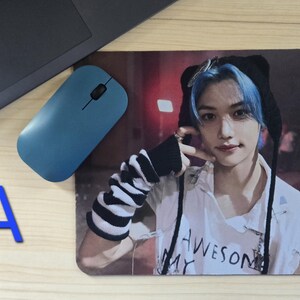 May include: A rectangular mousepad featuring a person with blue hair wearing a black hat and a white shirt with the text "AWESOME MY". A blue wireless mouse and a laptop are also visible.