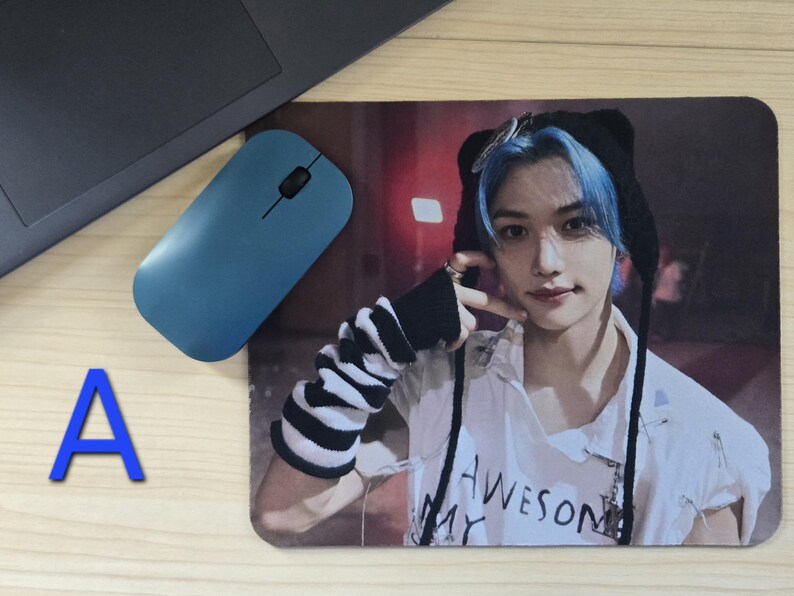 May include: A rectangular mousepad featuring a person with blue hair wearing a black hat and a white shirt with the text "AWESOME MY". A blue wireless mouse and a laptop are also visible.