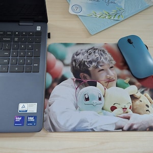 May include: A rectangular mousepad featuring a color photograph of a person holding three plush toys. A blue wireless mouse sits on the right. A laptop is on the left.