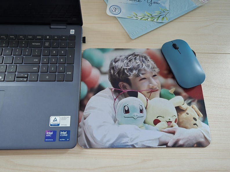 May include: A rectangular mousepad featuring a color photograph of a person holding three plush toys. A blue wireless mouse sits on the right. A laptop is on the left.