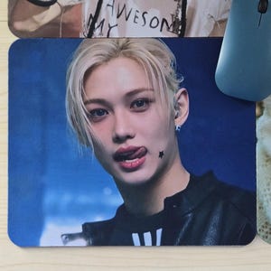 May include: A rectangular mousepad featuring a close-up image of a person with blonde hair and a black jacket. The person is sticking out their tongue. A blue computer mouse is on the right side of the mousepad.