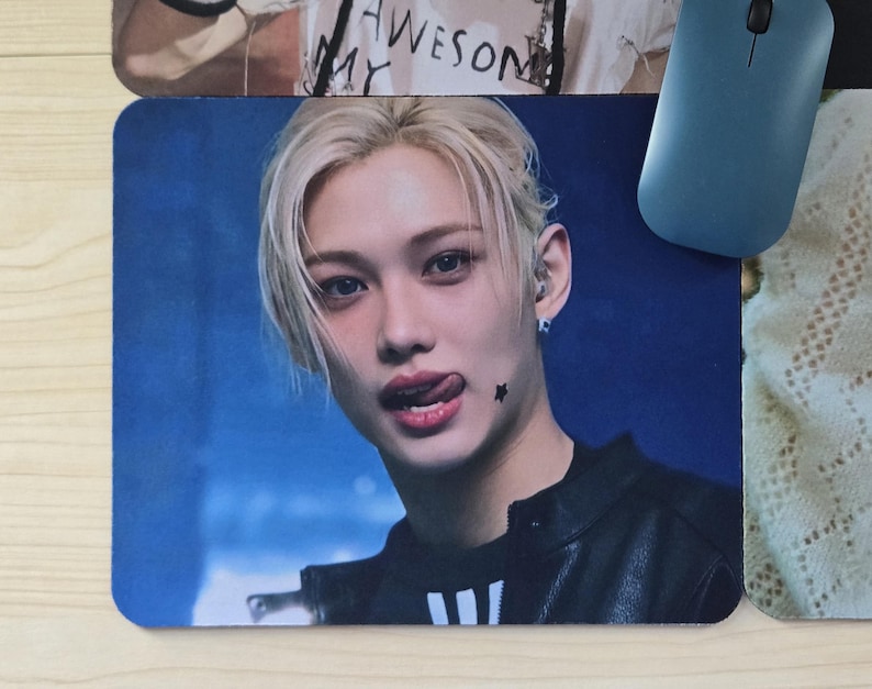 May include: A rectangular mousepad featuring a close-up image of a person with blonde hair and a black jacket. The person is sticking out their tongue. A blue computer mouse is on the right side of the mousepad.