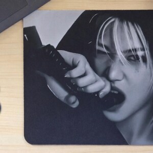 May include: Black and white mousepad featuring a person with white-streaked hair, biting a black object. The image is a close-up, with the person's face and hand in focus. The letter "B" is in blue in the lower left corner.