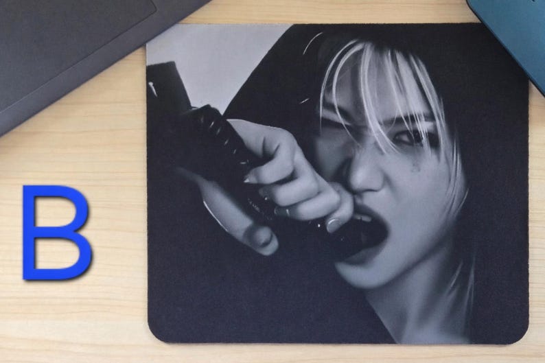 May include: Black and white mousepad featuring a person with white-streaked hair, biting a black object. The image is a close-up, with the person's face and hand in focus. The letter "B" is in blue in the lower left corner.