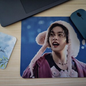 May include: A mousepad with a person wearing a pink hat with ear-like flaps. The person is wearing a pink jacket and a black necklace. A blue computer mouse and a card with the words "Thank You" are also visible.