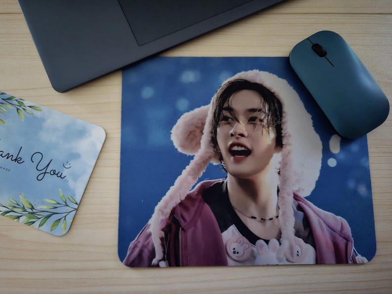 May include: A mousepad with a person wearing a pink hat with ear-like flaps. The person is wearing a pink jacket and a black necklace. A blue computer mouse and a card with the words "Thank You" are also visible.