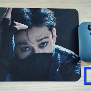 May include: A rectangular mousepad featuring a close-up image of a person with dark hair and striking blue eyes. A blue wireless mouse is positioned to the right. A blue letter "D" is in the lower right corner.