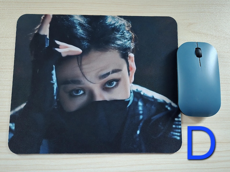 May include: A rectangular mousepad featuring a close-up image of a person with dark hair and striking blue eyes. A blue wireless mouse is positioned to the right. A blue letter "D" is in the lower right corner.