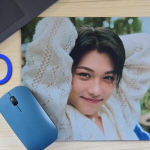 May include: A rectangular mousepad featuring a smiling person with dark hair wearing a white sweater. A blue computer mouse and the letter "D" are visible. The mousepad is on a light-colored surface.