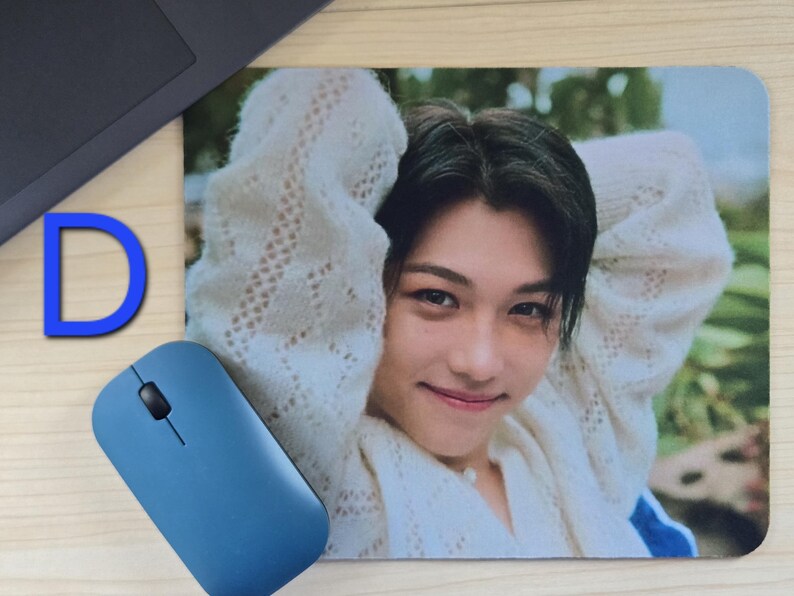 May include: A rectangular mousepad featuring a smiling person with dark hair wearing a white sweater. A blue computer mouse and the letter "D" are visible. The mousepad is on a light-colored surface.