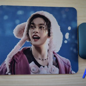 May include: Rectangular mouse mat featuring a person wearing a pink hooded hat, a pink jacket, and a black shirt. A blue wireless mouse is to the right of the mouse mat. The letter "A" is in blue.