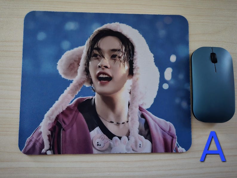 May include: Rectangular mouse mat featuring a person wearing a pink hooded hat, a pink jacket, and a black shirt. A blue wireless mouse is to the right of the mouse mat. The letter "A" is in blue.