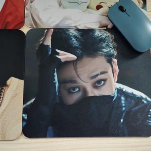 May include: A black mousepad featuring a close-up of a person with dark hair and striking blue eyes. The person's face is partially obscured by a black garment. A blue computer mouse rests on the surface.