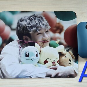May include: A rectangular mousepad featuring a person holding plush toys of cartoon characters. A blue wireless mouse is to the right. The letter "A" is in the lower right corner. The background has a blurred image of balloons.