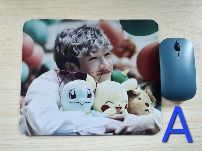 May include: A rectangular mousepad featuring a person holding plush toys of cartoon characters. A blue wireless mouse is to the right. The letter "A" is in the lower right corner. The background has a blurred image of balloons.
