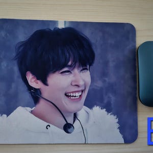 May include: A rectangular mouse mat featuring a smiling person with dark hair and a white hoodie. A teal wireless mouse is positioned to the right. The letter "B" is visible in blue.