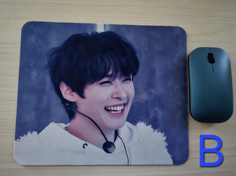 May include: A rectangular mouse mat featuring a smiling person with dark hair and a white hoodie. A teal wireless mouse is positioned to the right. The letter "B" is visible in blue.