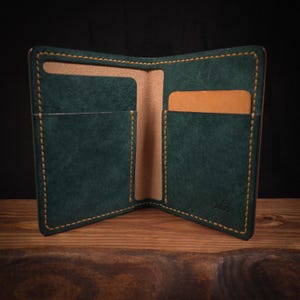 May include: Open, dark green leather wallet with tan interior and stitching. The wallet has multiple card slots and a bill compartment. The initials "MA" are visible on the lower right corner.