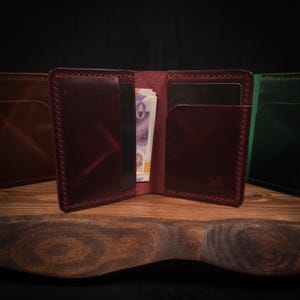 May include: Open burgundy leather wallet with multiple card slots and cash. The wallet is displayed open, revealing the interior and contents. Other wallets in brown and green are visible in the background.