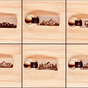 May include: A collection of six keychains with mountain landscape designs. Each keychain features a rectangular tag with a mountain scene etched in brown, attached to a metal ring and a leather strap.