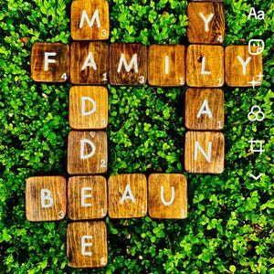 Könnte beinhalten: Holz-Scrabble-Steine buchstabieren "FAMILY" und "DAD BEAU" in Kreuzform, vor dem Hintergrund von grünem Laub. Die Steine sind braun mit weißen Buchstaben und Zahlen.