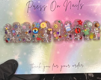 May include: Handmade press-on nails with a rainbow glitter base and various colorful 3D embellishments, including a smiley face, an eye, a donut, a flower, and an M. The text "Handmade Press On Nails" is written on the card.