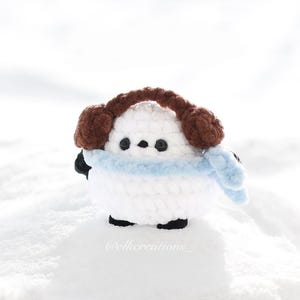 Può includere: Un pupazzo di neve fatto a mano all'uncinetto con corpo bianco, piedi neri e una sciarpa azzurra. Il pupazzo di neve indossa paraorecchie marroni. La figura è ambientata su uno sfondo innevato.