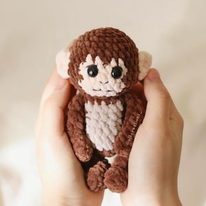 以下が含まれることがあります： 茶色とクリーム色の手作りの編み物モンキー人形。 ぬいぐるみは、黒い目と笑顔の表情が特徴です。 猿は両手で持たれています。
