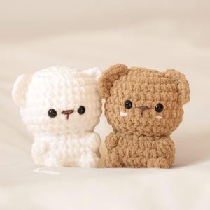 May include: Two handmade crochet bear toys. One is white, the other is brown. Both have black eyes and small brown noses. The bears are sitting side by side on a white surface. The toys are soft and cuddly.